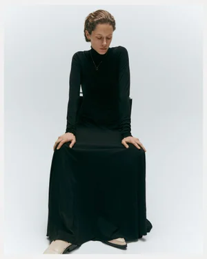 Model seated wearing a long black turtleneck dress against a clean white studio background.