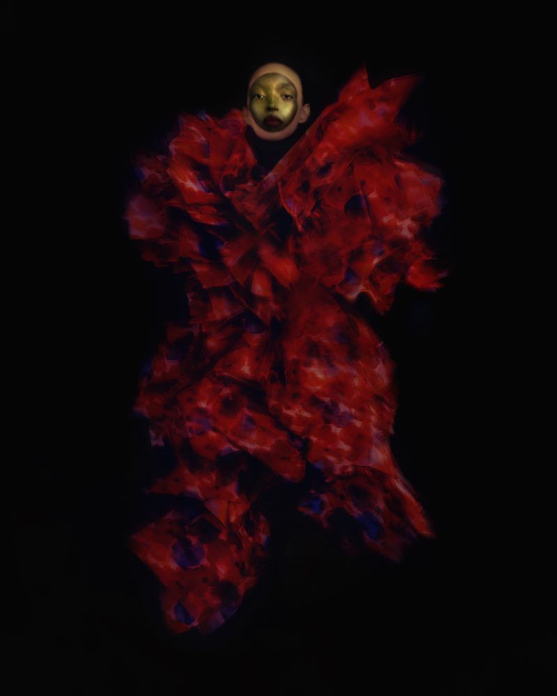 A figure with a gold-painted face emerging from darkness wearing an expansive, textured red ruffled garment.