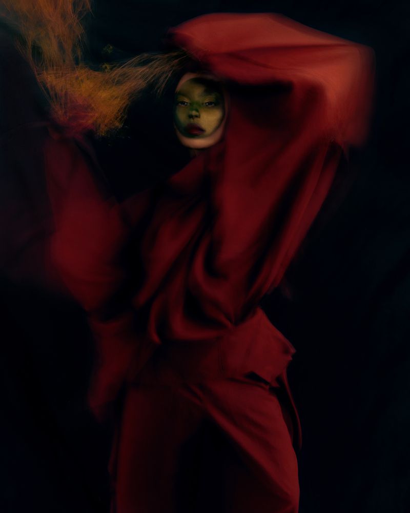 Editorial portrait of a model wrapped in dynamic flowing crimson fabric against a dark background with artistic makeup.