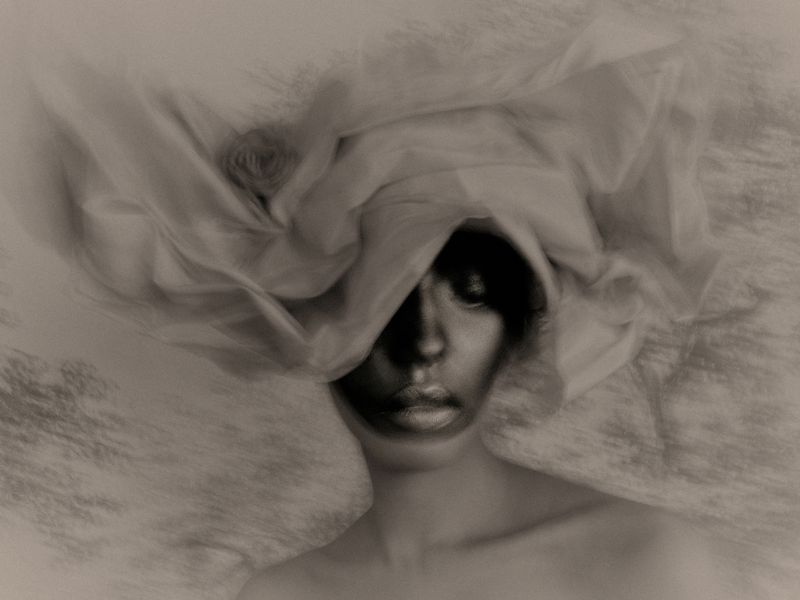 Close-up portrait of a woman wearing an oversized draped fabric headpiece with dramatic shadows across her face.