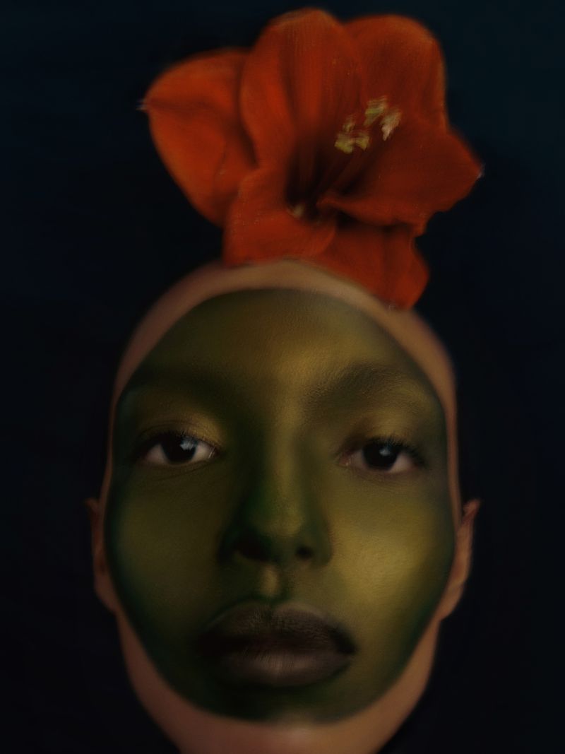 Close-up portrait of a person with green face paint and a large red flower on their head against a black background.