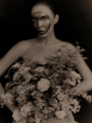 Monochrome portrait of a woman holding a large cluster of flowers against a dark studio background in soft focus.