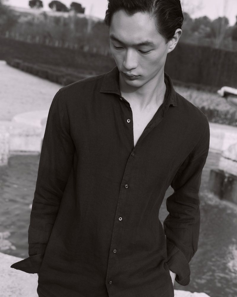 East Asian man looking down wearing a black unbuttoned long-sleeve shirt outdoors by a stone fountain.