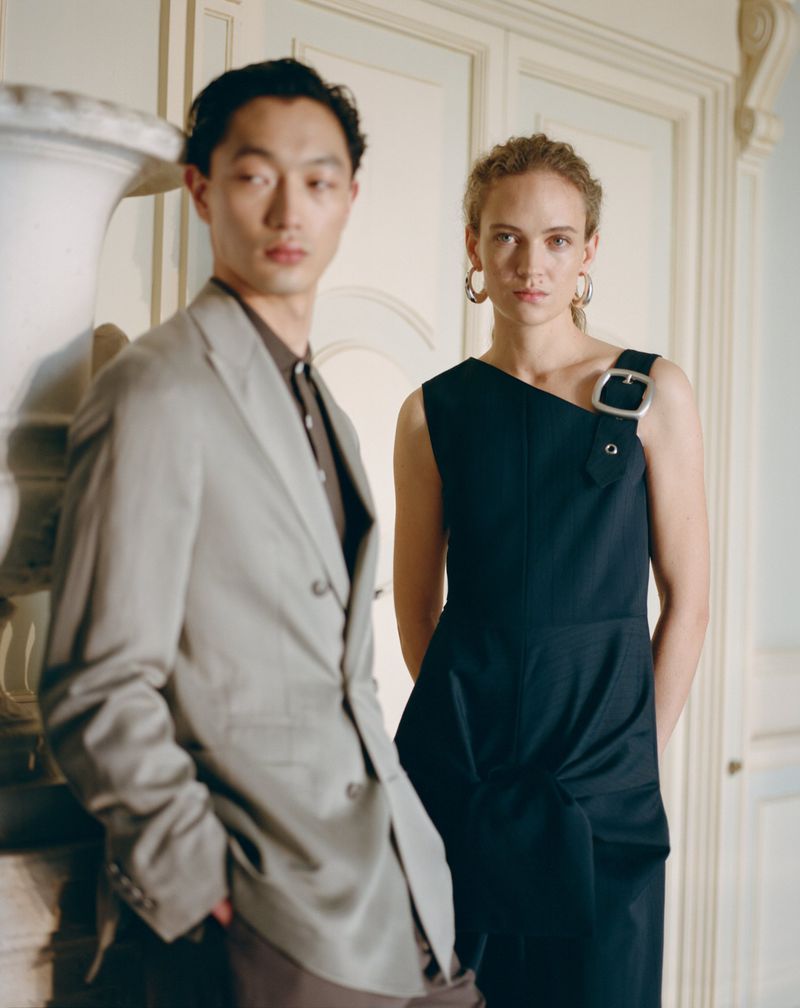 East Asian man in a grey suit and woman in a dark sleeveless dress with an oversized shoulder buckle posing in an elegant room.