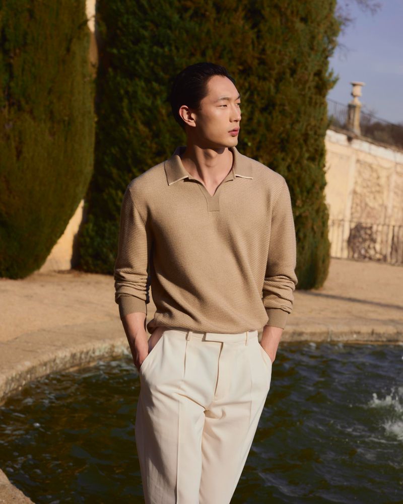 East Asian man wearing a beige knit polo shirt and cream pleated trousers standing beside an outdoor fountain pool in sunlight.