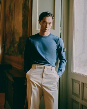 East Asian man in a blue long-sleeve knit sweater and cream tailored trousers leaning against a window frame.