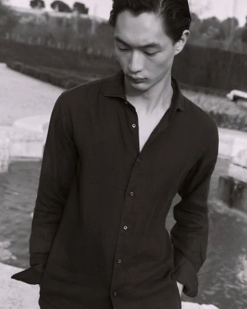 East Asian man looking down wearing a black unbuttoned long-sleeve shirt outdoors by a stone fountain.