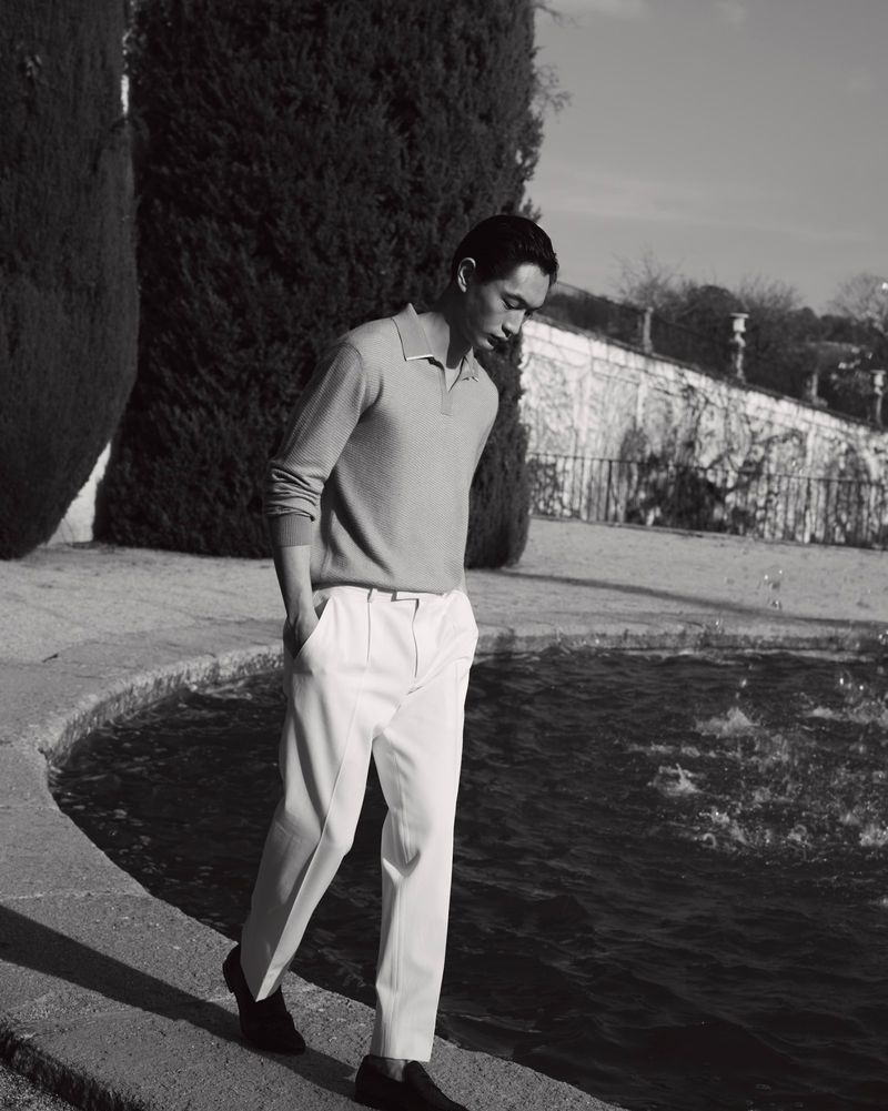 East Asian man in a long-sleeve polo shirt and pleated white trousers walking along the edge of an outdoor stone fountain.