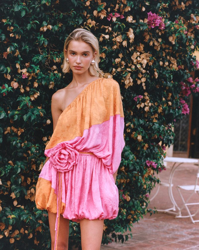 Model in a one-shoulder pink and orange mini dress with rosette detail posing in front of a blooming bougainvillea hedge.
