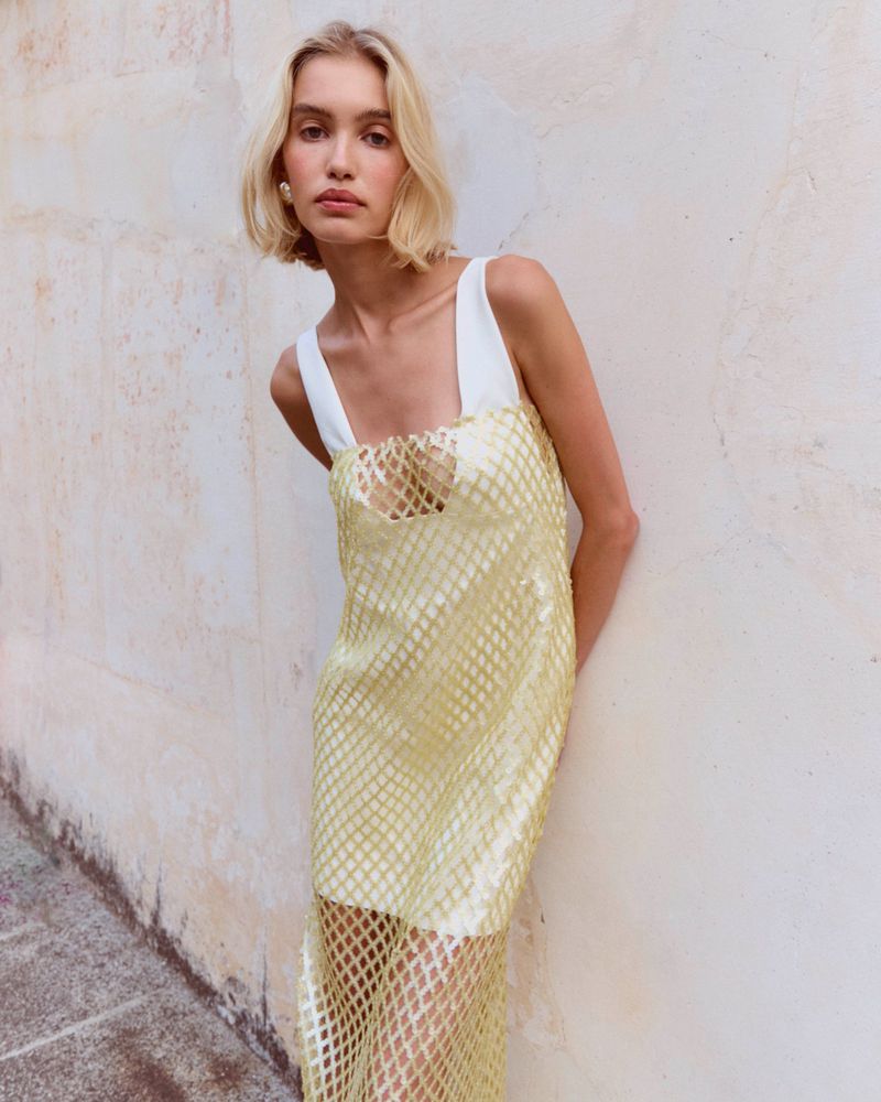 Blonde model in a pale yellow netted overlay dress leaning against a textured wall.