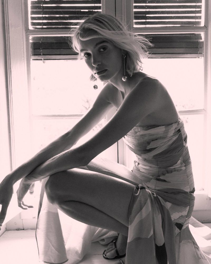 Blonde woman in a strapless patterned dress crouching beside an interior window with slatted blinds.