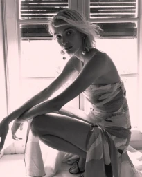 Blonde woman in a strapless patterned dress crouching beside an interior window with slatted blinds.