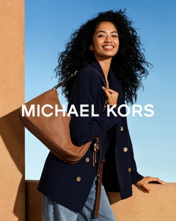 Smiling woman with dark curly hair wearing a navy blazer and holding a brown shoulder bag against a blue sky.