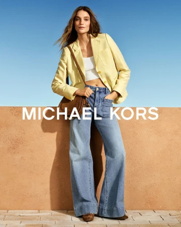 Model wearing a pale yellow blazer and wide-leg denim jeans leaning against a terracotta wall under a clear blue sky.