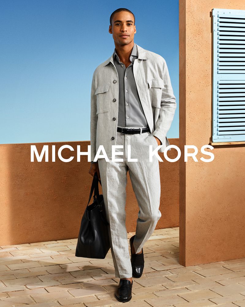 Model Malik Lindo wearing a grey linen jacket and trousers while carrying a black leather bag on a sunlit terracotta terrace.