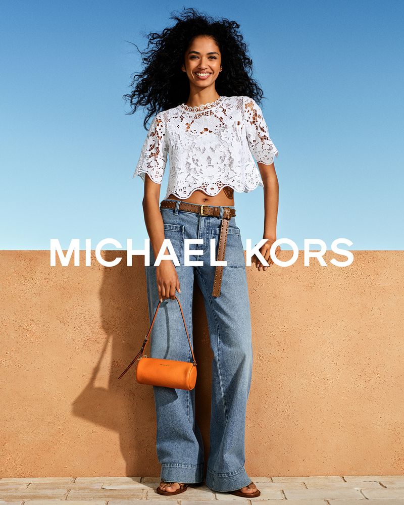 A woman poses with a tan bag, wearing a white lace top and denim pants against a backdrop of blue sky and beige stone.