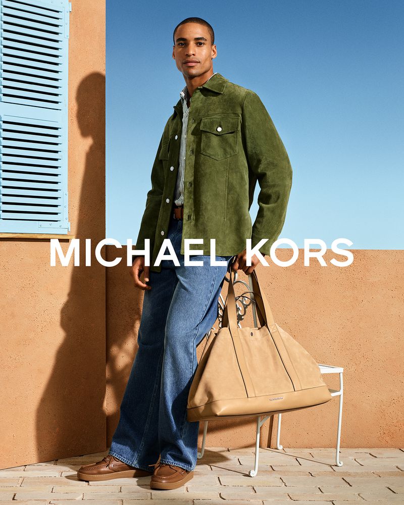 Malik Lindo in an olive green suede jacket and blue denim jeans holding a large beige Michael Kors tote bag outdoors.