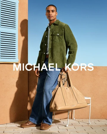 Malik Lindo in an olive green suede jacket and blue denim jeans holding a large beige Michael Kors tote bag outdoors.
