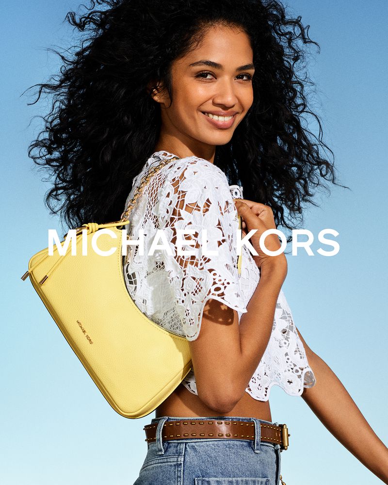 Smiling model with voluminous curly hair wearing a white lace top and carrying a pastel yellow shoulder bag against a blue sky.