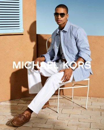 Model Malik Lindo sits on a white chair wearing a blue striped shirt, white trousers, and sunglasses against a warm stucco wall.