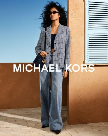 Model posing against a terracotta stucco wall with blue shutters wearing a plaid blazer, wide-leg jeans, and sunglasses for Michael Kors.