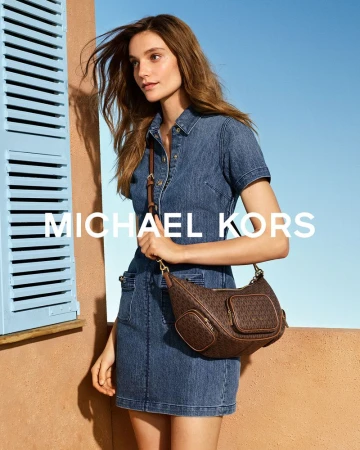 Model wearing a short-sleeved blue denim dress and brown monogram Michael Kors crossbody bag by a terracotta wall with blue shutters.