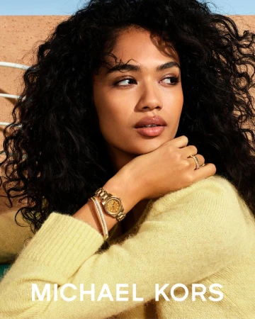 Close-up portrait of a woman wearing a pale yellow sweater, gold watch, and bracelet against an outdoor textured background.