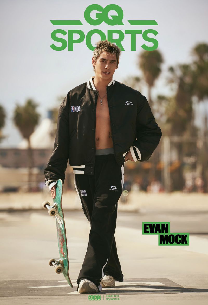 Evan Mock walking outdoors holding a skateboard while wearing an open black bomber jacket and matching track pants.