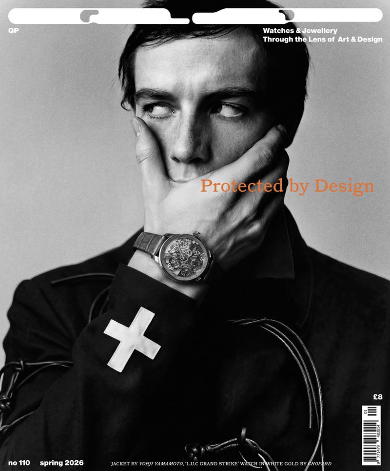 Man covering his mouth with a luxury skeleton watch visible on his wrist, wearing a black jacket with a white cross patch.