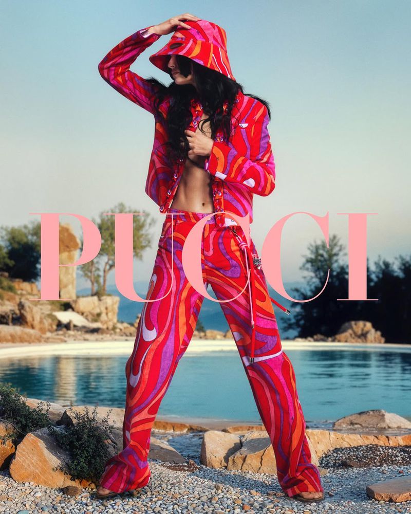 Fashion shot of a woman wearing a bright patterned jacket, pants, and hat, standing outdoors with a swimming pool and landscape background.