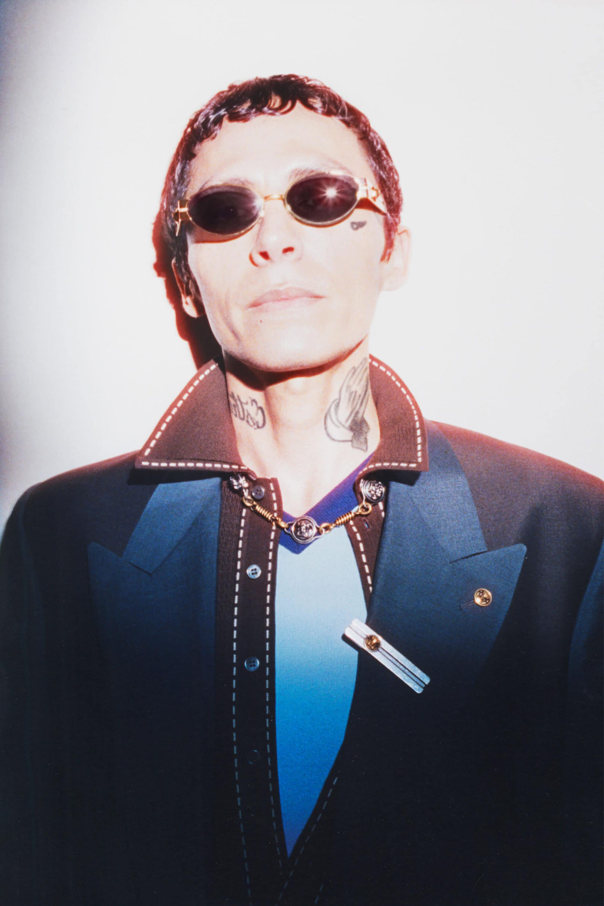 Chuquimamani Condori wears dark oval sunglasses, a brown collared shirt, and a tailored black blazer against a light background.
