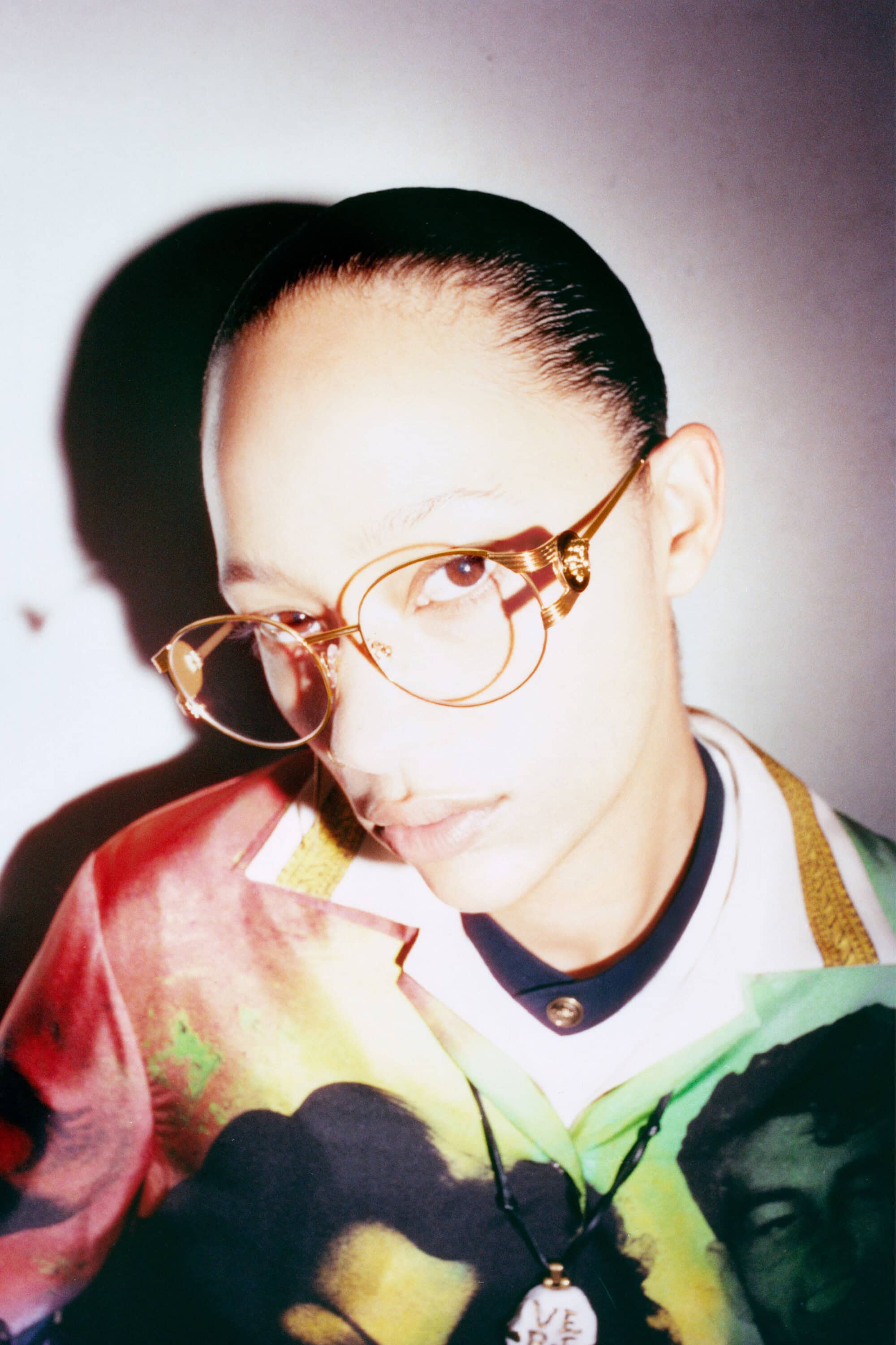 Model Selena Forrest wearing sculptural gold wire eyeglasses and a colorful graphic collared shirt against a light backdrop.