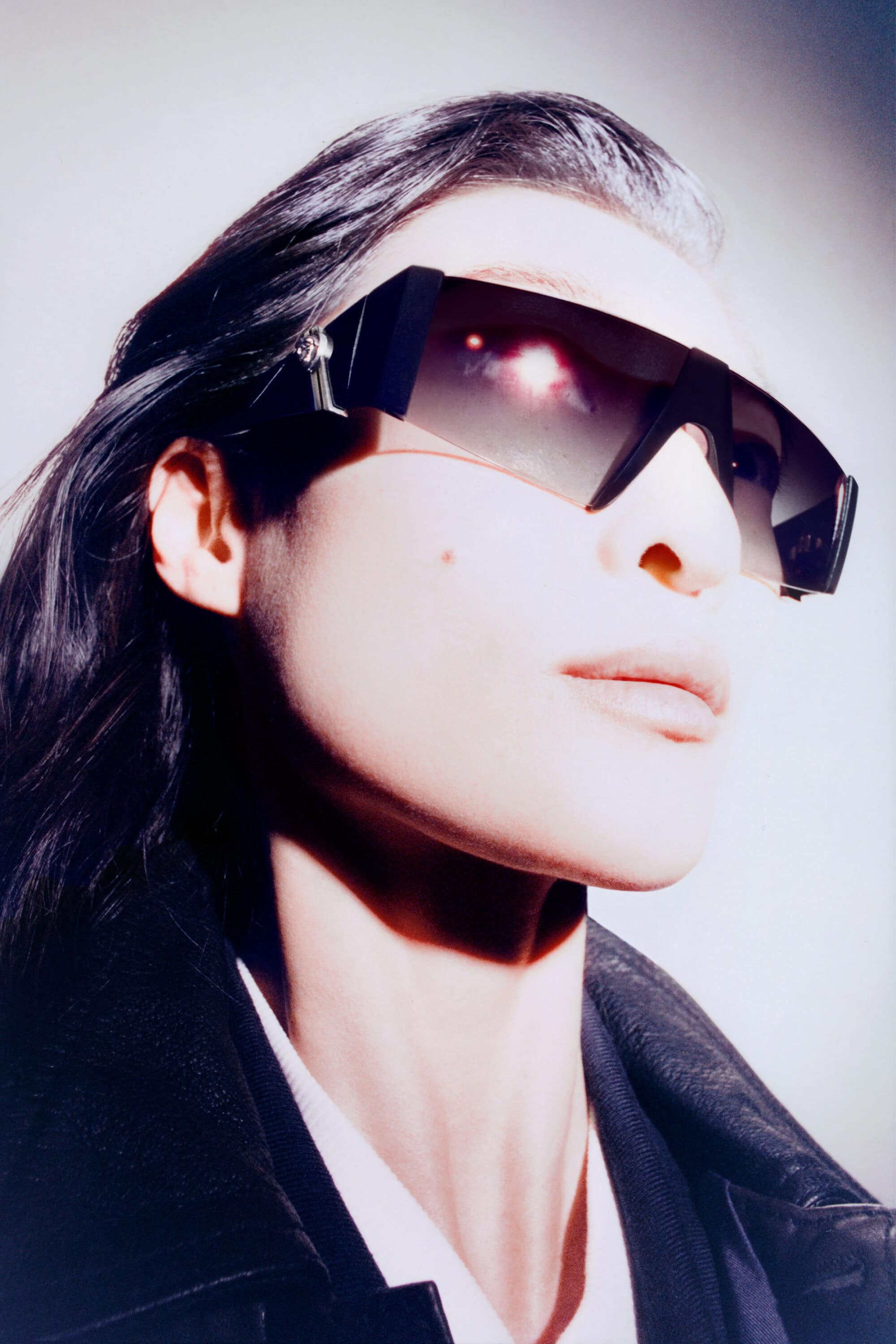 Close-up portrait of a model wearing bold black shield sunglasses and a dark collared jacket against a neutral studio backdrop.