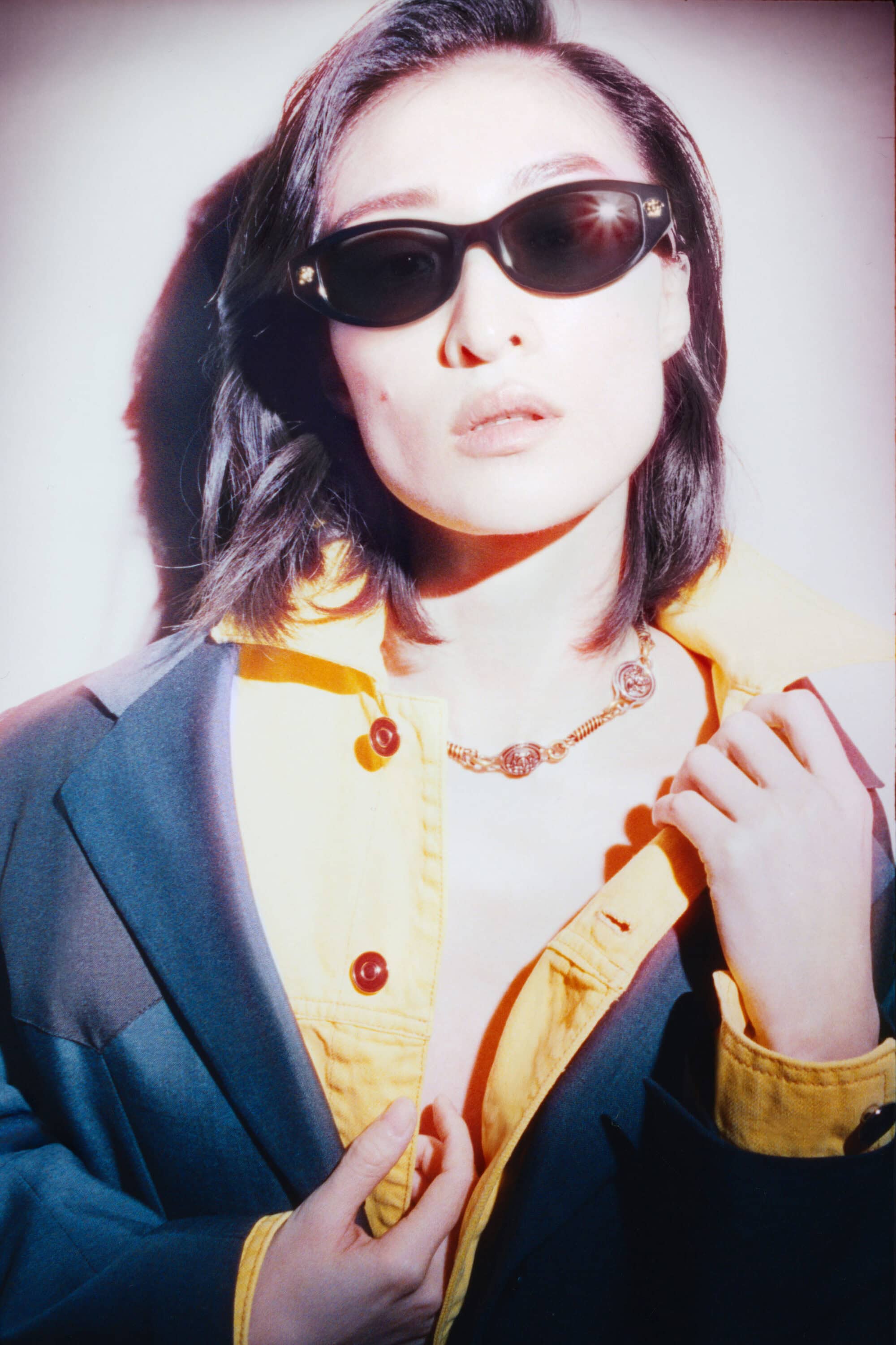 Model wearing dark sunglasses, a yellow shirt, and dark jacket against a plain wall for Versace eyewear campaign.