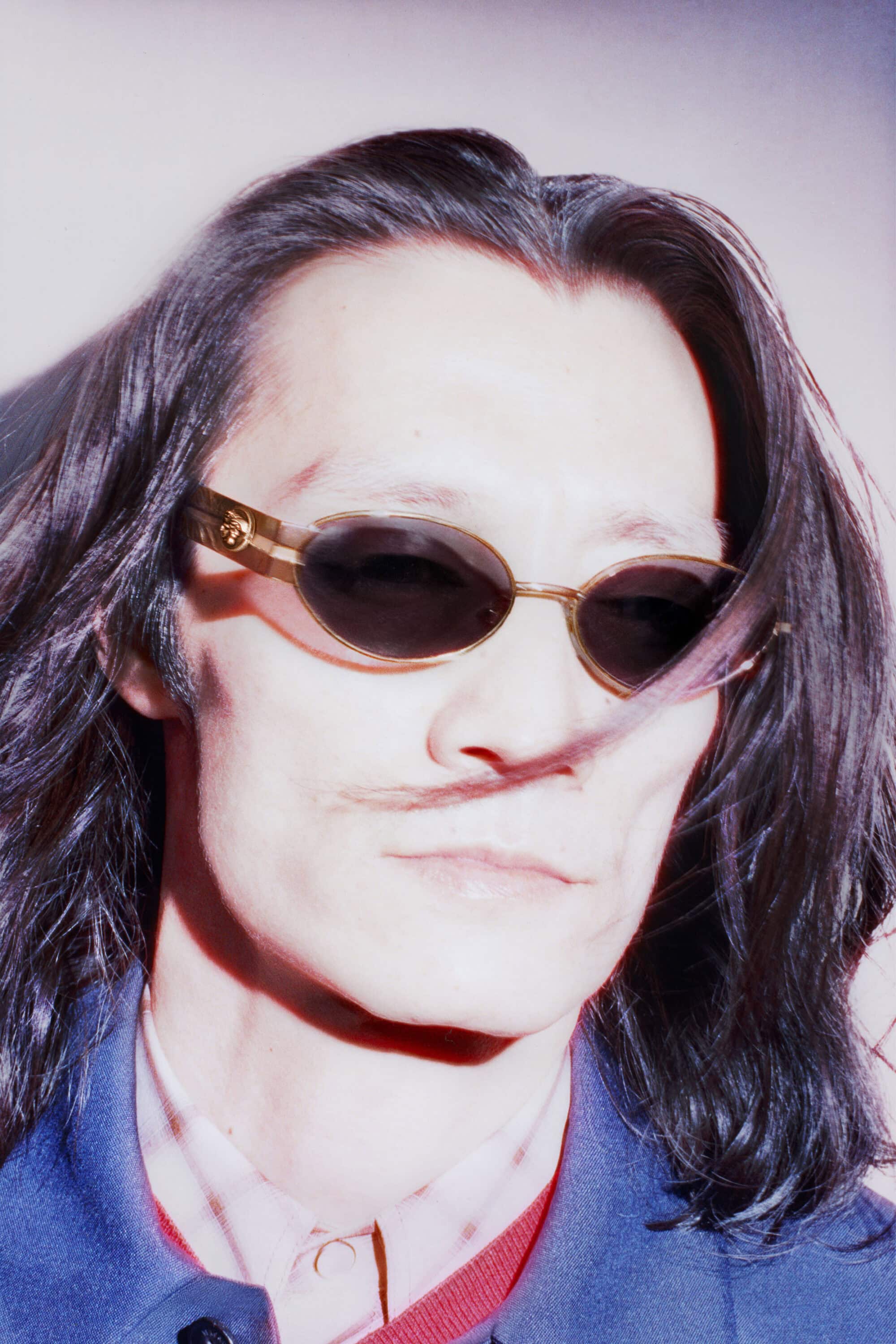 Kazuya Tanabe wearing gold oval sunglasses and a blue jacket in a close-up portrait for the Versace Eyewear campaign.