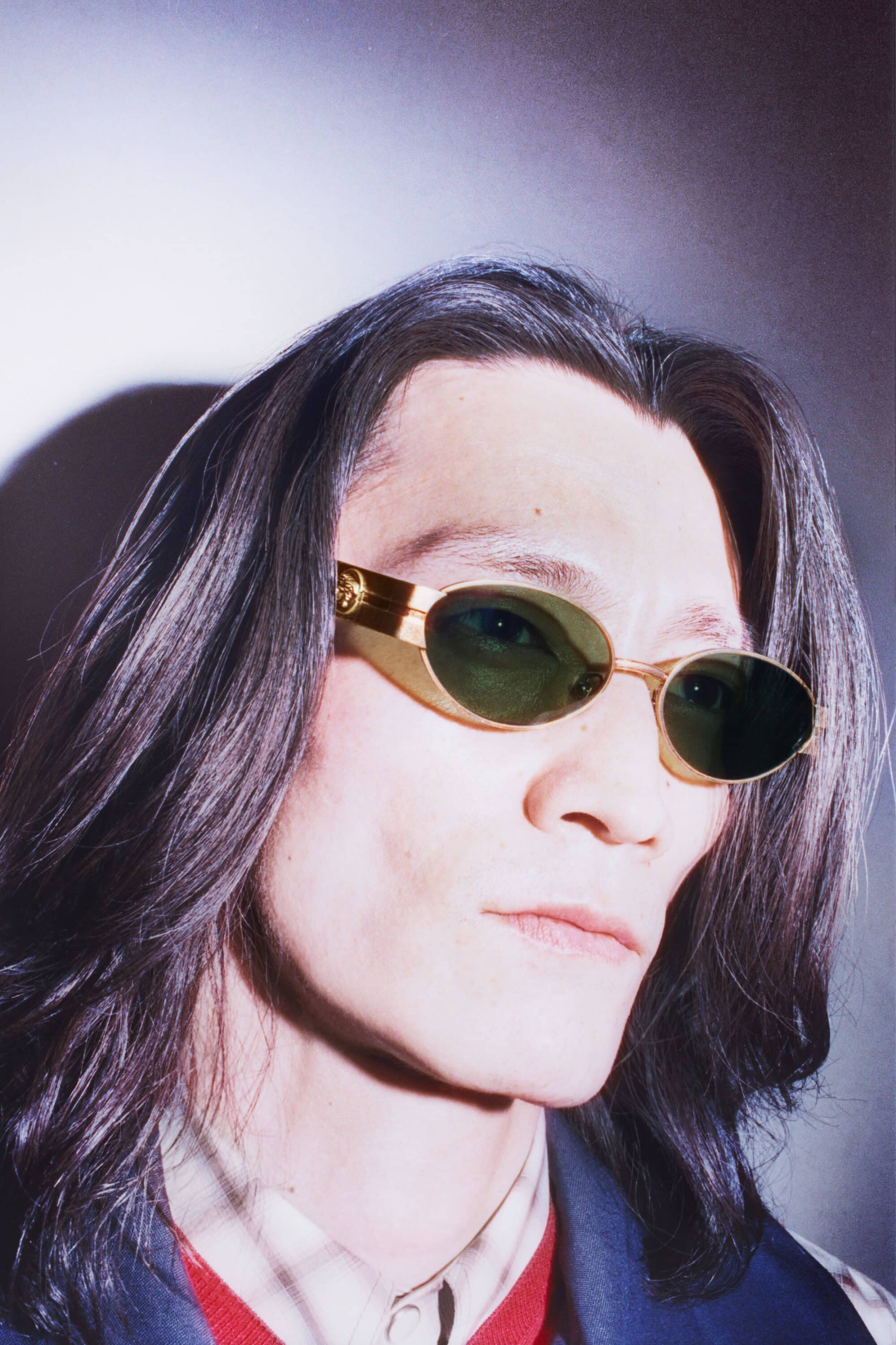 Portrait of a person with long hair wearing gold sunglasses, standing indoors against a mottled light and dark background.