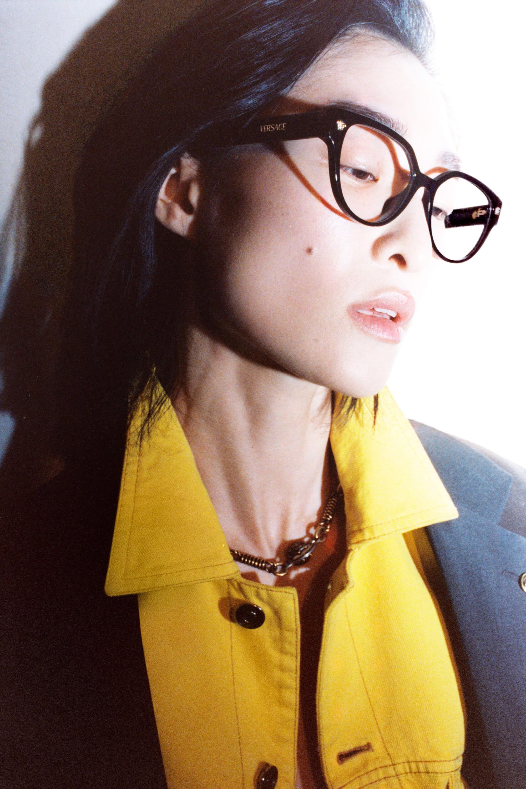 Chu Wong wears black Versace eyeglasses and a bright yellow collared jacket lit with sharp direct shadows.