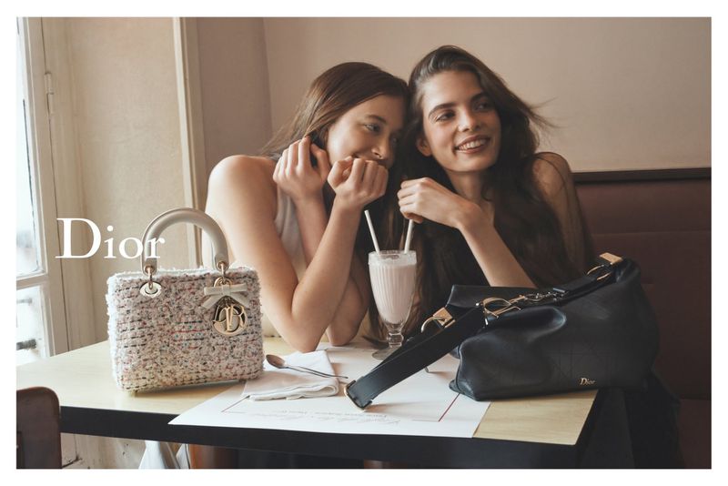 Two young women sitting closely at a cafe table with a textured Lady Dior handbag, black shoulder bag, and a milkshake.
