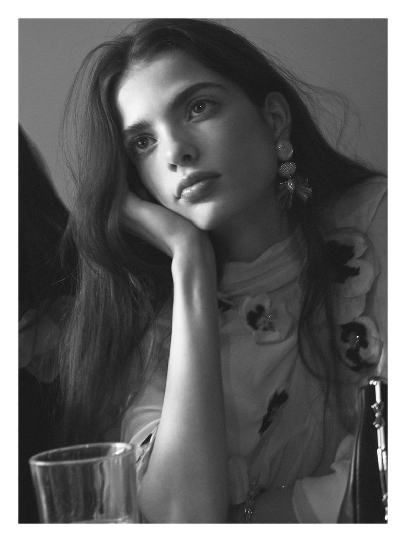 Black and white portrait of a woman resting her chin on her hand, wearing statement drop earrings and a floral textured top.