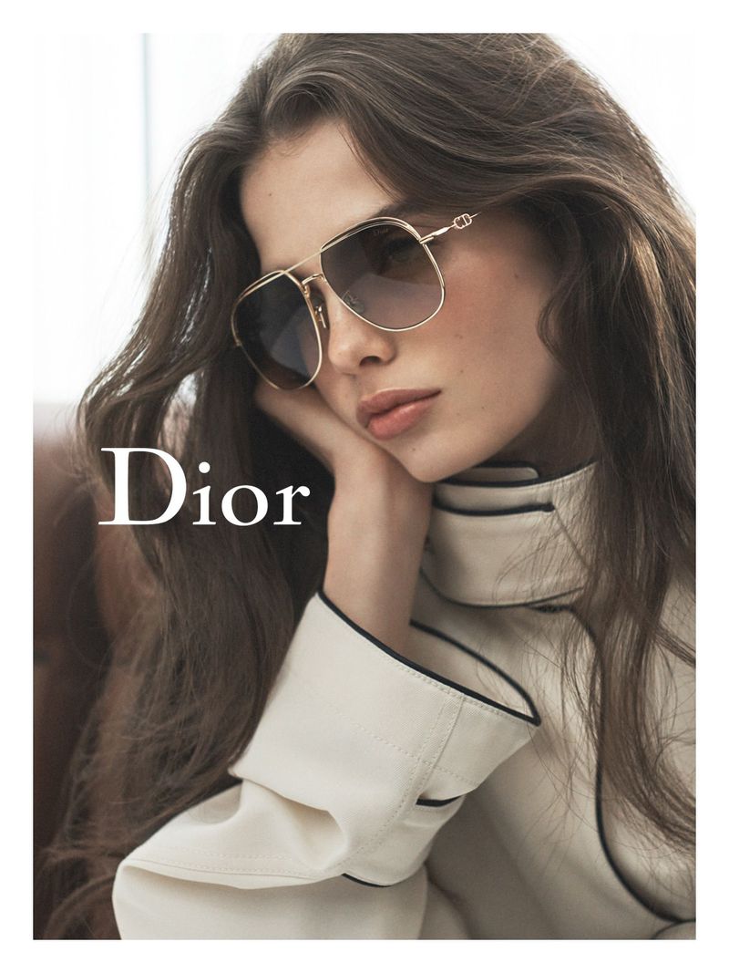 Model wearing gold aviator sunglasses with gradient lenses and a cream collared jacket with black piping beside a white Dior logo.