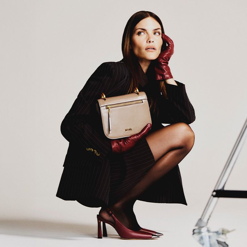 Fashion shot of a woman in a pinstripe suit and leather gloves holding a beige handbag, posing against a neutral backdrop in a studio.