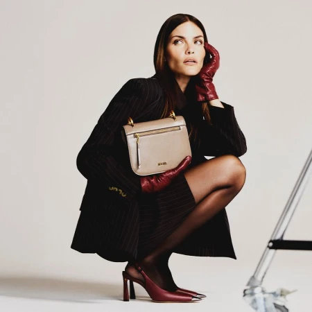 Fashion shot of a woman in a pinstripe suit and leather gloves holding a beige handbag, posing against a neutral backdrop in a studio.