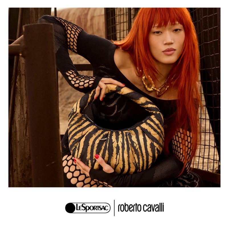 A redhead woman poses with a black and brown zebra-striped LeSportsac x Roberto Cavalli shoulder bag in an indoor setting.