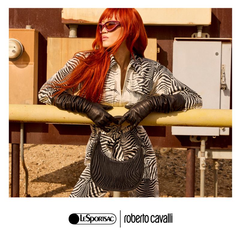 Fashion shot of a redhead woman in zebra-print outfit, long leather gloves, sunglasses, and purse, posing outdoors with metal background.