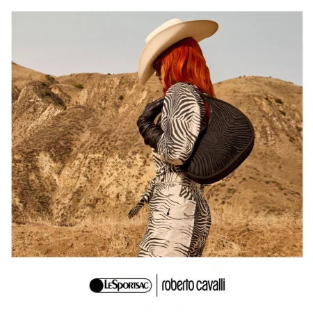 A woman in a zebra-print dress and cowboy hat poses in a desert landscape with a black purse in a fashion shot.