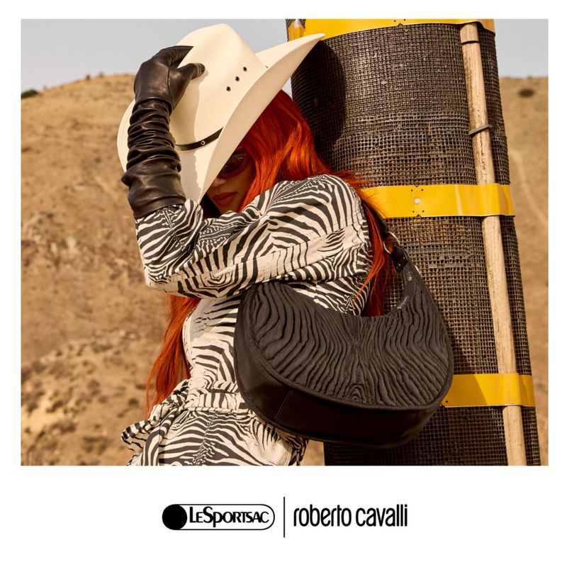 A person in a zebra-patterned outfit with a black purse and white cowboy hat, standing near a pole in a desert environment.