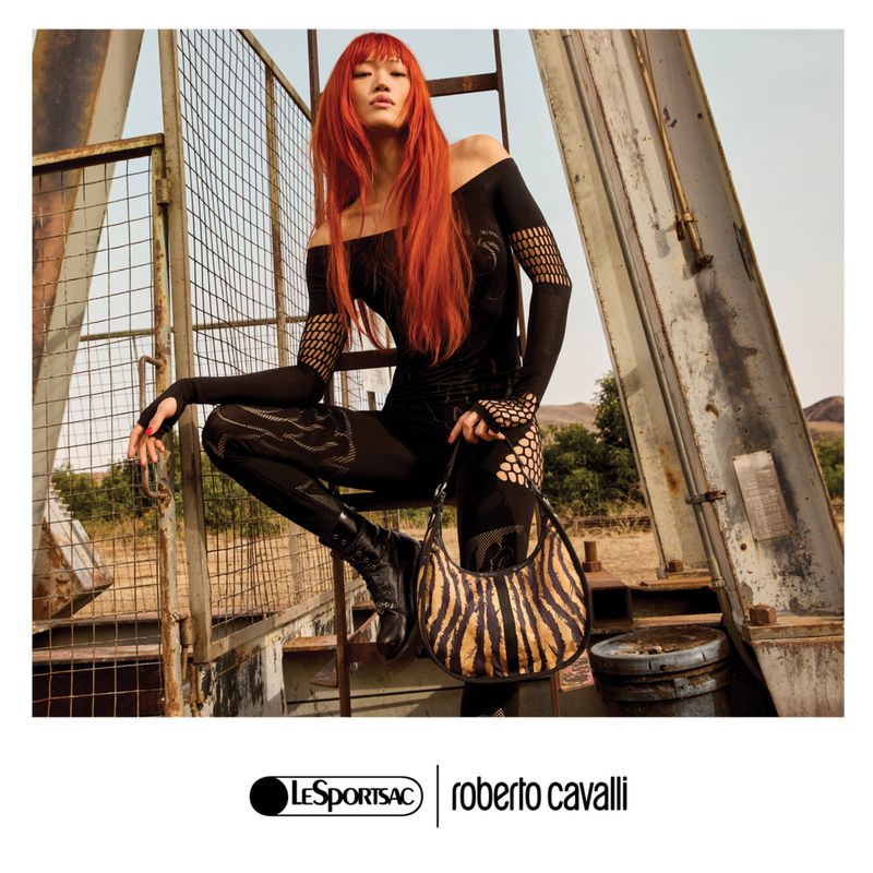 Fashion shot of an adult woman with red hair wearing a black catsuit and boots, holding a tiger-striped bag, posing on a weathered metal structure in an outdoor setting.
