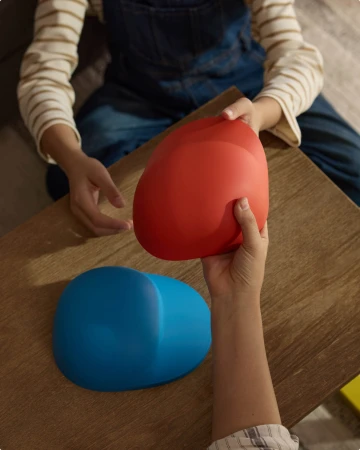 High angle view of hands passing a smooth matte red curved object over a wooden table near a matching blue object.