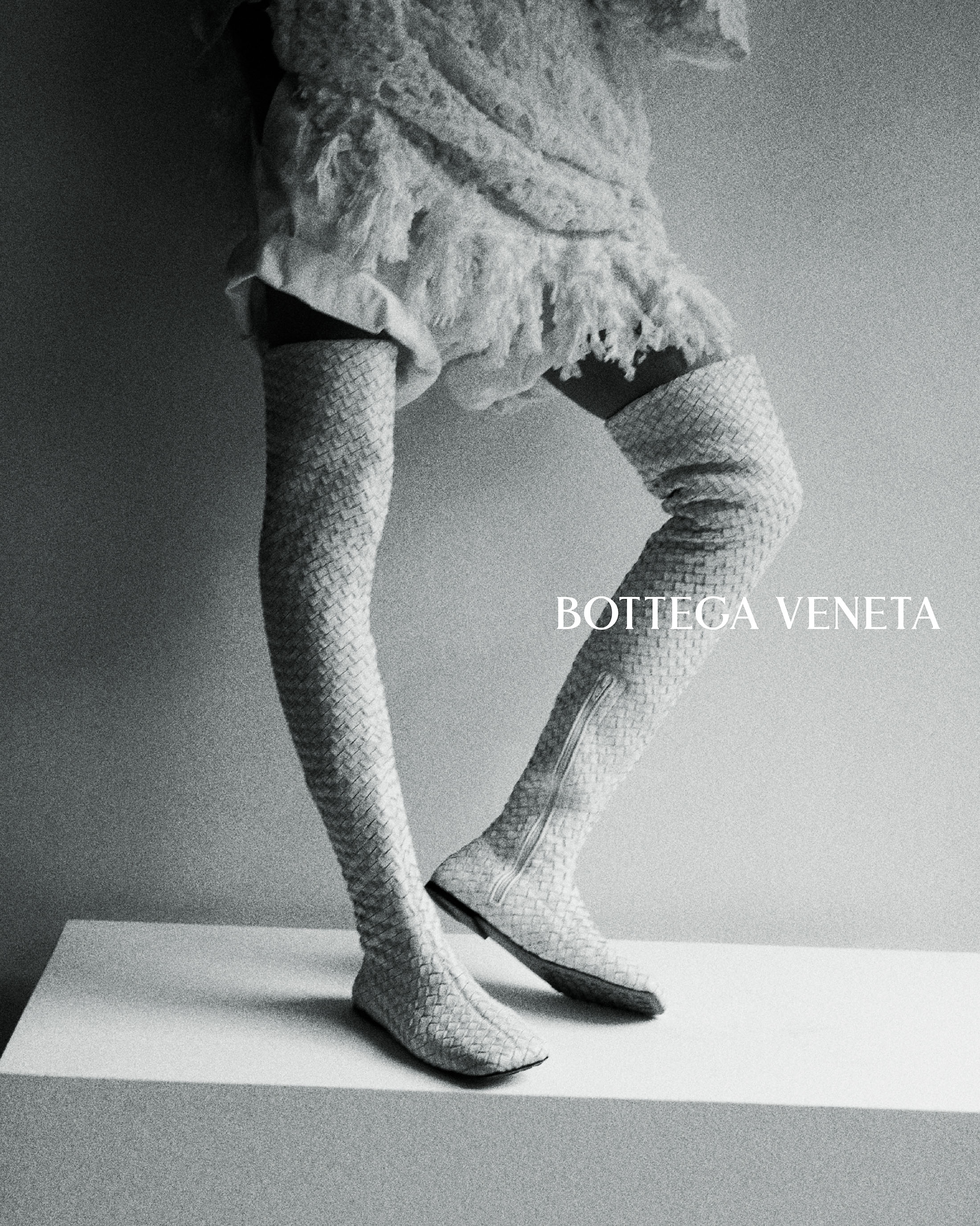 Model wearing textured fringe dress and woven thigh-high leather boots standing on a white plinth in a Bottega Veneta campaign.