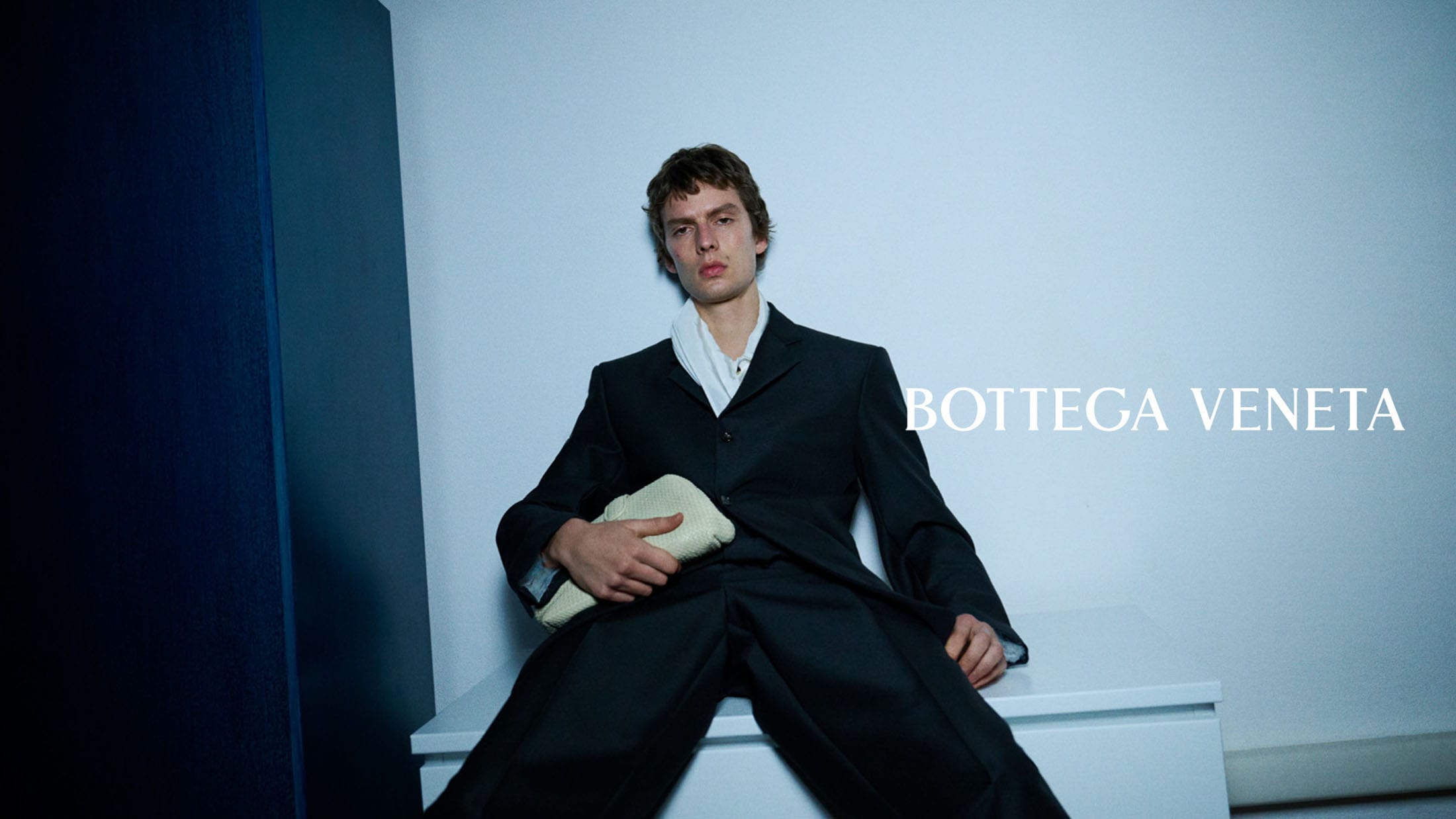 Leon Dame in a black tailored suit seated with a woven clutch bag in a Bottega Veneta campaign